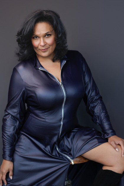 Maya Francine Thomas seated in a navy zip dress with a sly half-smile