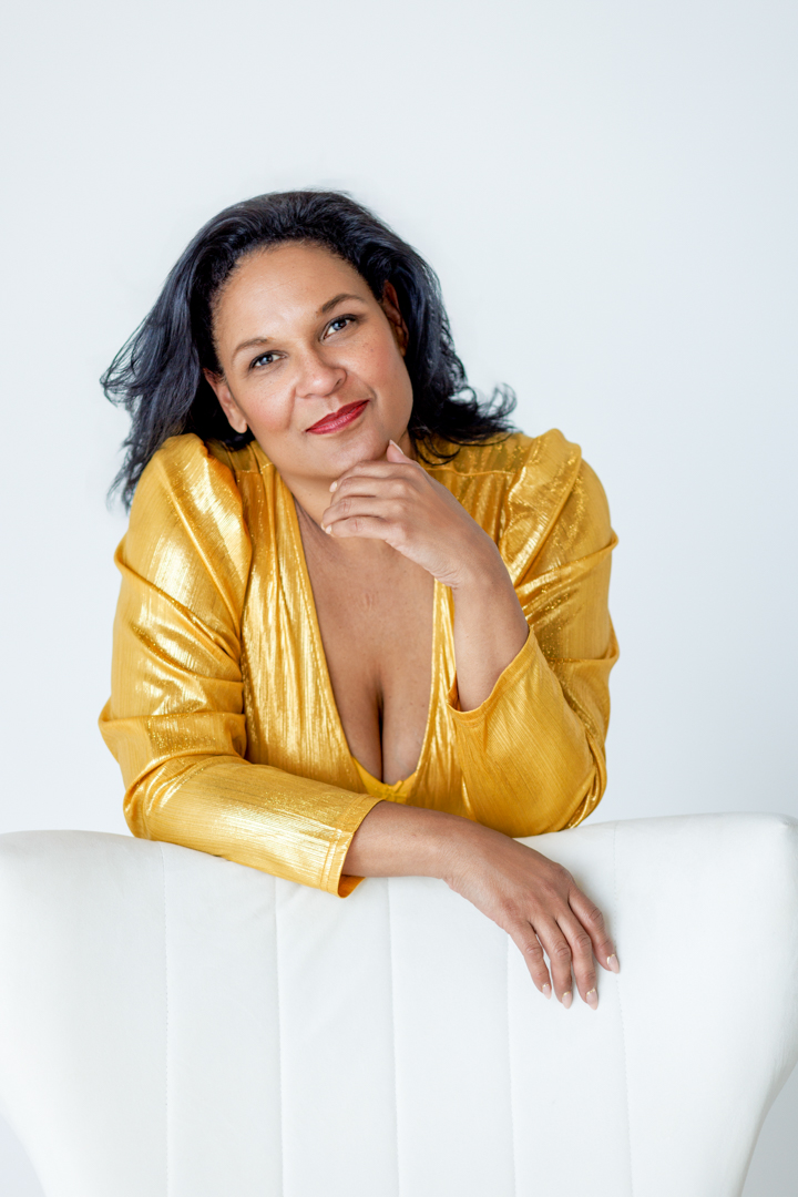 Maya Francine Thomas in gold, leaning on a white chair